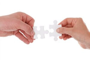 Two hands each holding a puzzle piece, bringing them together to connect.